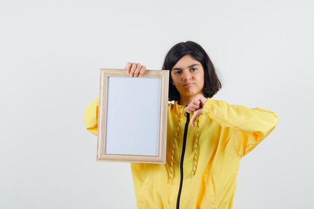 Het jonge whiteboard van de meisjesholding, die duim in geel bomberjack toont en ontevreden kijkt