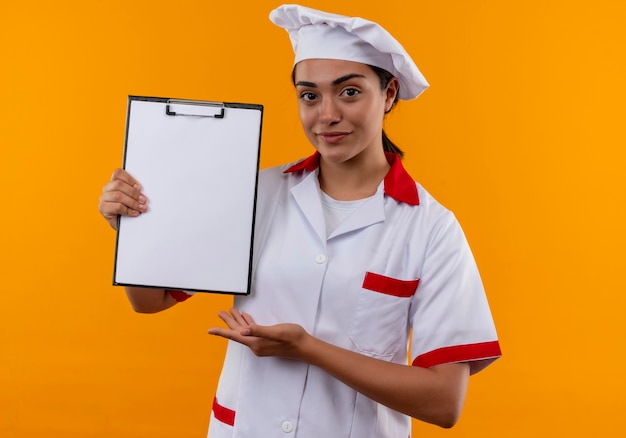 Het jonge zekere Kaukasisch kokmeisje in eenvormige chef-kok houdt klembord en punten met hand die op oranje muur met exemplaarruimte wordt geïsoleerd