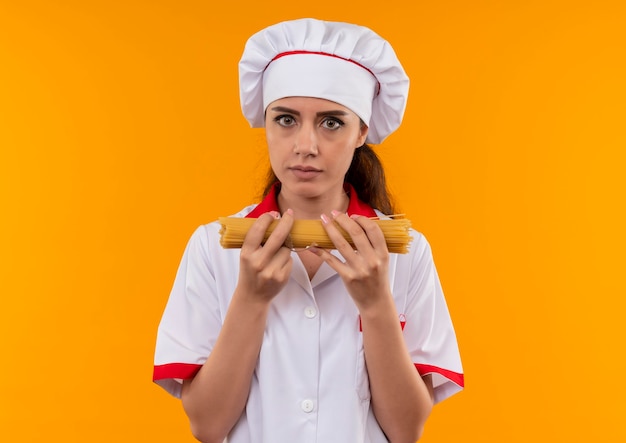 Het jonge zelfverzekerde Kaukasisch kokmeisje in eenvormige chef-kok houdt bos van spaghetti met beide handen die op oranje muur met exemplaarruimte wordt geïsoleerd