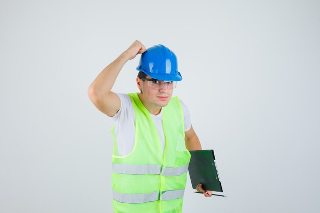 Het klembord en de pen van de jongensholding, hand op helm in bouwuniform zetten en ernstig, vooraanzicht kijken.