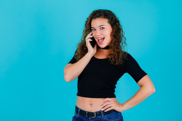 Het lachende meisje praat over de telefoon door de hand op de taille op een blauwe achtergrond te leggen