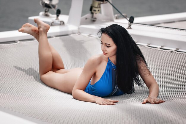 Het langharige volwassen meisje in blauwe strakke bikini ontspant op het net van het privéjacht in de zee, portret
