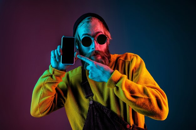 Het lege scherm van de telefoon wordt weergegeven. Kaukasisch man's portret op de achtergrond van de gradiëntstudio in neonlicht. Mooi mannelijk model met hipsterstijl. Concept van menselijke emoties, gezichtsuitdrukking, verkoop, advertentie.