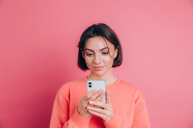 Het leuke gelukkige jonge mooie vrouw stellen geïsoleerd over roze muurachtergrond die mobiele telefoon met behulp van