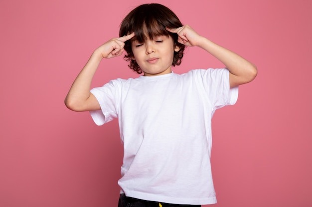 Het leuke schattige portret van de kindjongen in wit t-shirt en zwarte broeken op roze bureau