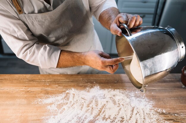 Het mannelijke bakkers gieten kneedt deeg van container op houten die lijst met bloem wordt bestrooid