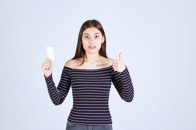 Het meisje in gestreept overhemd houdt een adreskaartje en toont duim omhoog teken.