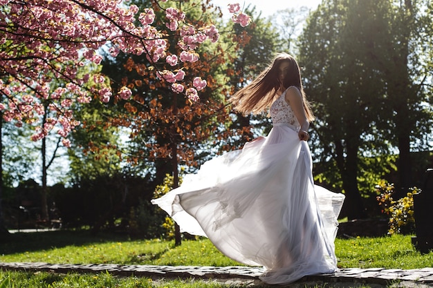 Het meisje in witte kleding bevindt zich onder roze bloeiende sakuraboom in het park