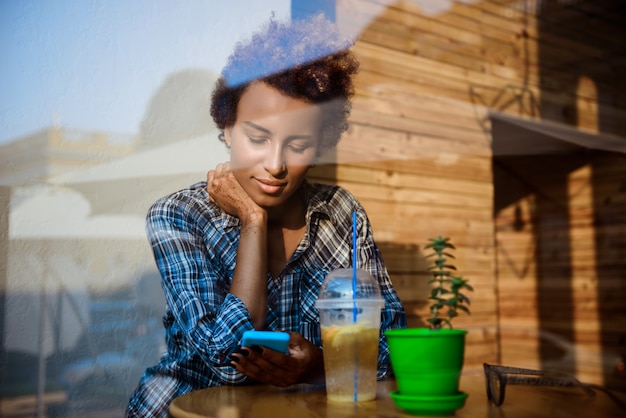 Het mooie Afrikaanse meisje glimlachen, die telefoon bekijken, die in koffie zitten. Schot van buitenaf.