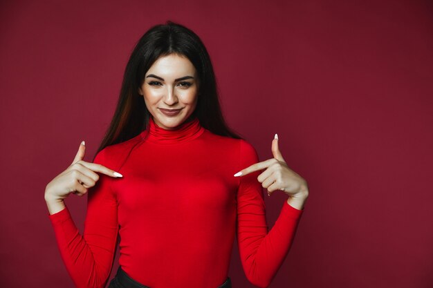 Het mooie geglimlachte donkerbruine Kaukasische meisje gekleed in rode trui toont haar borsten
