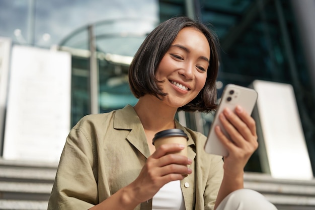 Het mooie glimlachende aziatische meisje dat koffie drinkt met mobiele telefoon en zit op de trap buiten de jonge wom