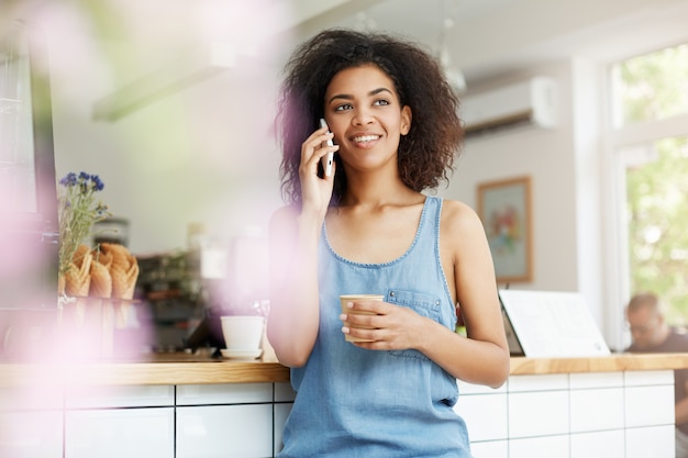 Het mooie vrolijke jonge Afrikaanse studente glimlachen die op telefoon het drinken koffie in koffie spreken.