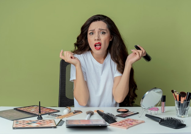 Het onaangename jonge mooie meisje zit aan make-up tafel met make-up tools houden kam en houden een andere hand in lucht geïsoleerd op olijfgroene achtergrond