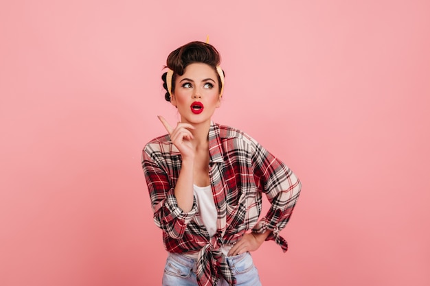 Het peinzende pinupmeisje stellen op roze achtergrond. Studio shot van charmante brunette vrouw, gekleed in geruit overhemd.