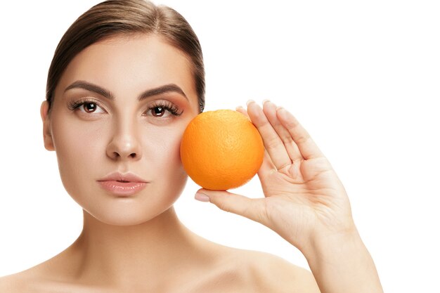 Het portret van aantrekkelijke Kaukasische glimlachende vrouw die op witte muur met oranje fruit wordt geïsoleerd. De schoonheid, verzorging, huid, behandeling, gezondheid, spa, cosmetica