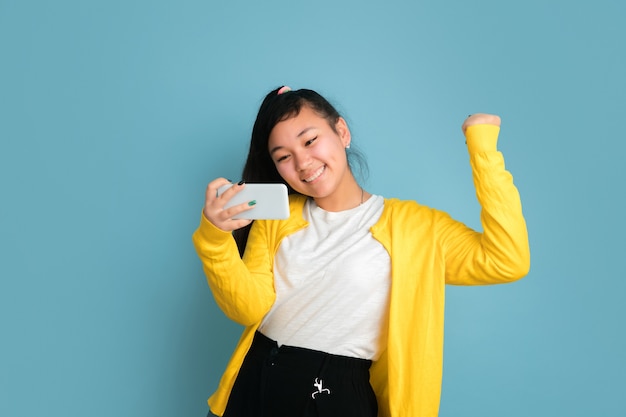 Het portret van de Aziatische tiener dat op blauwe studioachtergrond wordt geïsoleerd. Mooi vrouwelijk donkerbruin model met lang haar. Concept van menselijke emoties, gezichtsuitdrukking, verkoop, advertentie. Met behulp van telefoon, glimlachen, gelukkige overwinning.