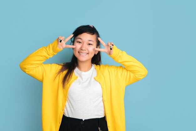 Het portret van de Aziatische tiener dat op blauwe studioachtergrond wordt geïsoleerd. Mooi vrouwelijk donkerbruin model met lang haar in informele stijl. Concept van menselijke emoties, gezichtsuitdrukking, verkoop, advertentie. Leuk poseren.