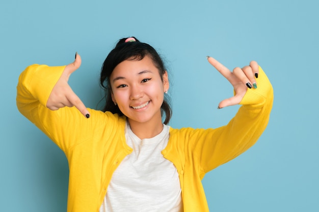 Het portret van de Aziatische tiener dat op blauwe studioachtergrond wordt geïsoleerd. Mooi vrouwelijk donkerbruin model met lang haar in informele stijl. Concept van menselijke emoties, gezichtsuitdrukking, verkoop, advertentie. Maakt selfie.