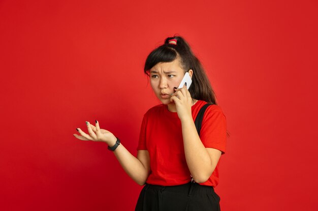 Het portret van de Aziatische tiener dat op rode studioachtergrond wordt geïsoleerd. Mooi vrouwelijk donkerbruin model met lang haar in informele stijl. Concept van menselijke emoties, gezichtsuitdrukking, verkoop, advertentie. Praten aan de telefoon.