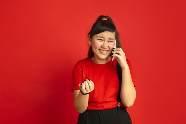 Het portret van de Aziatische tiener dat op rode studioachtergrond wordt geïsoleerd. Mooi vrouwelijk donkerbruin model met lang haar in informele stijl. Concept van menselijke emoties, gezichtsuitdrukking, verkoop, advertentie. Praten aan de telefoon.