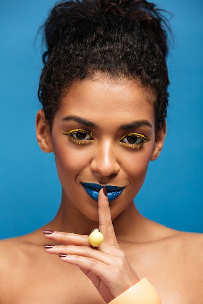 Het portret van de close-upschoonheid van halfnaakte afrikaanse vrouw met maniermake-up die stilte of geheim vragen die vinger op lippen geïsoleerd, over blauwe muur vragen te houden