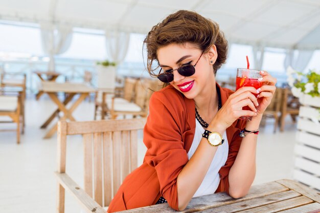 Het portret van de de zomerlevensstijl van het leuke vrij jonge vrouw openlucht stellen, zittend in strandkoffie en het drinken van exotische cocktail, overzeese achtergrond. Felle kleuren. Vakantie stemming. Glimlachen en plezier hebben.