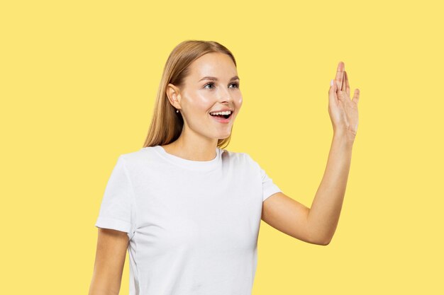 Het portret van de halve lengte van de kaukasische jonge vrouw op gele studioachtergrond. mooi vrouwelijk model in wit overhemd. concept van menselijke emoties, gezichtsuitdrukking. groet, iemand uitnodigen.