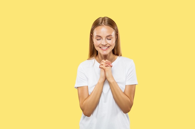 Het portret van de halve lengte van de Kaukasische jonge vrouw op gele studioachtergrond. Mooi vrouwelijk model in wit overhemd. Concept van menselijke emoties, gezichtsuitdrukking, verkoop. Bidden met gesloten ogen, glimlachend.