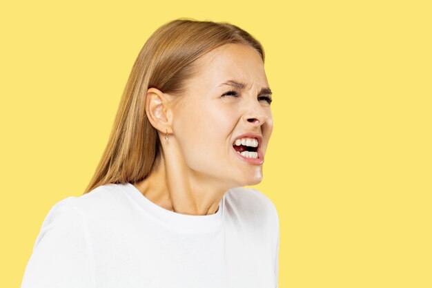 Het portret van de halve lengte van de Kaukasische jonge vrouw op gele studioachtergrond. Mooi vrouwelijk model in wit overhemd. Concept van menselijke emoties, gezichtsuitdrukking, verkoop. Boos, boos, agressief geschreeuw.