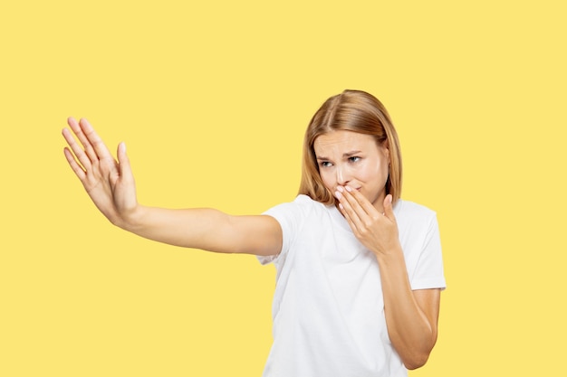 Het portret van de halve lengte van de Kaukasische jonge vrouw op gele studioachtergrond. Mooi vrouwelijk model in wit overhemd. Concept van menselijke emoties, gezichtsuitdrukking. Ziet er walgelijk uit, van streek, bedekt haar gezicht.