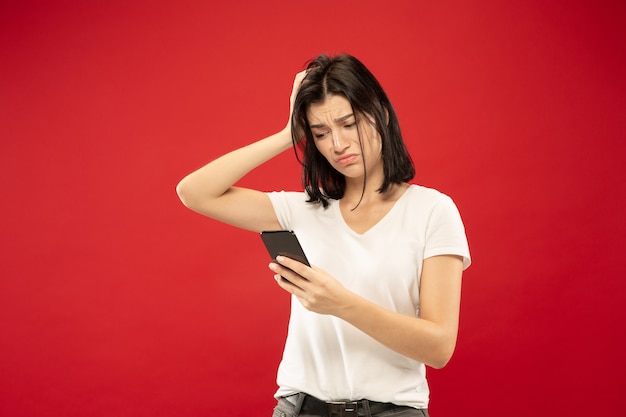 Het portret van de halve lengte van de Kaukasische jonge vrouw op rode studioachtergrond. Mooi vrouwelijk model in wit overhemd. Concept van menselijke emoties, gezichtsuitdrukking. Koopt online, betalingsrekening, ziet er boos uit.