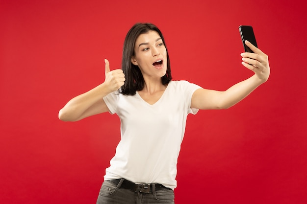 Het portret van de halve lengte van de Kaukasische jonge vrouw op rode studioachtergrond. Mooi vrouwelijk model in wit overhemd. Concept van menselijke emoties, gezichtsuitdrukking. Selfie maken of haar eigen vlog, blog.