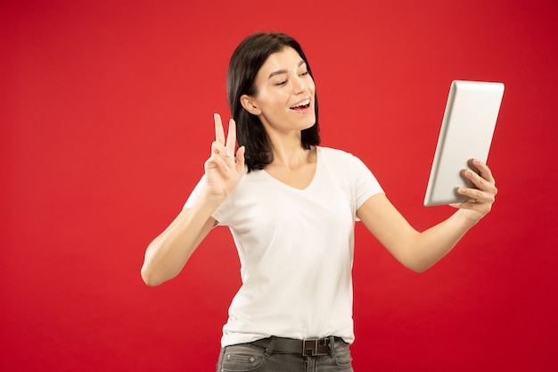 Het portret van de halve lengte van de Kaukasische jonge vrouw op rode studioachtergrond. Mooi vrouwelijk model in wit overhemd. Concept van menselijke emoties, gezichtsuitdrukking. Tablet gebruiken om haar vlog te maken of te praten.