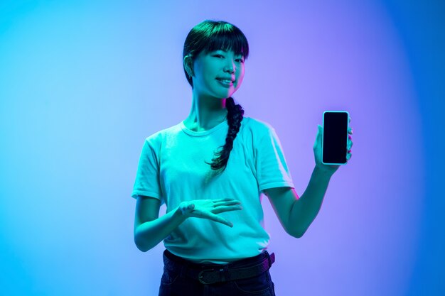 Het portret van de jonge Aziatische vrouw op achtergrond van de gradiënt de blauw-purpere studio in neonlicht. Concept van jeugd, menselijke emoties, gezichtsuitdrukking, verkoop, advertentie. Mooi donkerbruin model.