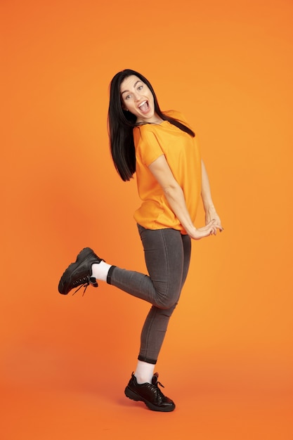 Het portret van de Kaukasische jonge vrouw op oranje studioachtergrond. Mooi vrouwelijk donkerbruin model in overhemd. Concept van menselijke emoties, gezichtsuitdrukking, verkoop, advertentie. Copyspace. Dansen, lachen.
