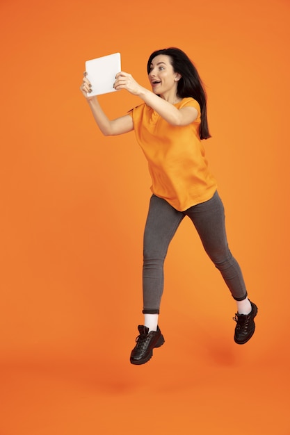 Het portret van de Kaukasische jonge vrouw op oranje studioachtergrond. Mooi vrouwelijk donkerbruin model in overhemd. Concept van menselijke emoties, gezichtsuitdrukking, verkoop, advertentie. Copyspace. Springen met tablet.