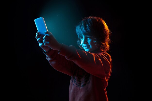 Het portret van de Kaukasische jongen dat op donkere studioachtergrond in neonlicht wordt geïsoleerd