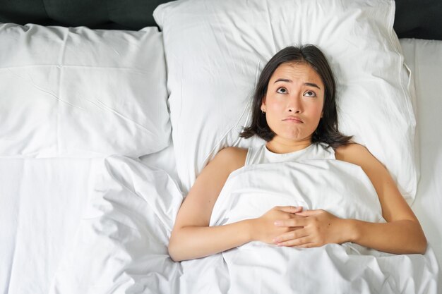 Het portret van een aziatisch meisje dat in haar bed ligt en haar schouders ophaalt, ziet er verbaasd uit voordat ze gaat slapen