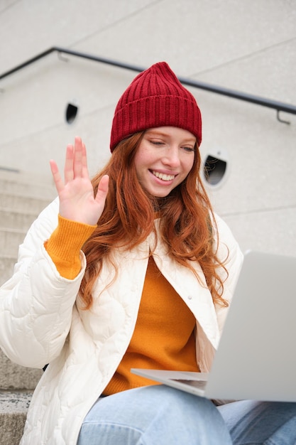 Het portret van een gelukkig jong roodharig meisje met laptop zwaait met de hand en zegt hallo op videochat heeft c