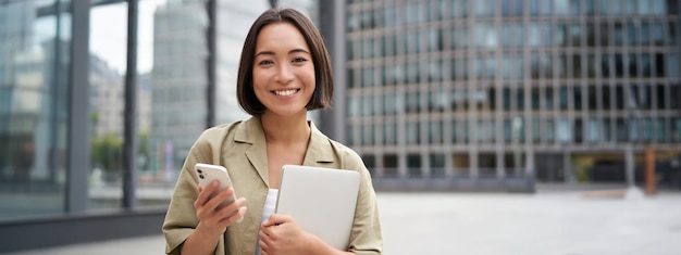 Gratis foto het portret van een glimlachend aziatisch meisje met een laptop houdt een mobiele telefoon vast en kijkt blij naar de camera die op