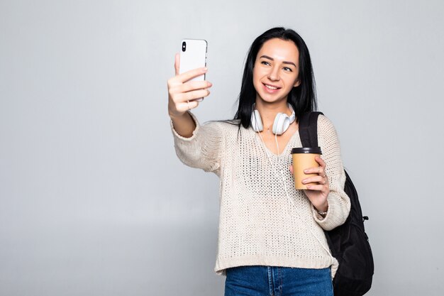 Het portret van een glimlachende aantrekkelijke vrouw die een selfie nemen terwijl het houden haalt koffiekop weg over witte muur wordt geïsoleerd die