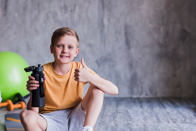 Het portret van een glimlachende jongenszitting met waterfles die duimen toont ondertekent omhoog