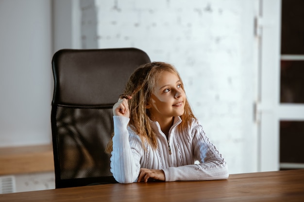 Het portret van een jong Kaukasisch meisje in vrijetijdskleding ziet er dromerig, schattig en gelukkig uit. Opzoeken en nadenken, binnen aan de houten tafel zitten. Concept van toekomst, doel, dromen, visualisatie.