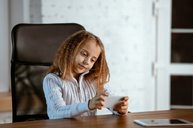 Het portret van een jong Kaukasisch meisje ziet er dromerig, schattig en gelukkig uit. Opzoeken, binnen zitten aan de houten tafel met tablet en smartphone. Concept van toekomst, doel, droom om te kopen, visualisatie.