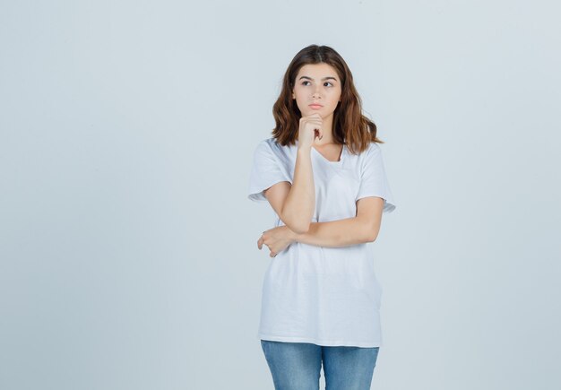 Het portret van een jong meisje steunt kin op hand in wit t-shirt en kijkt peinzend vooraanzicht