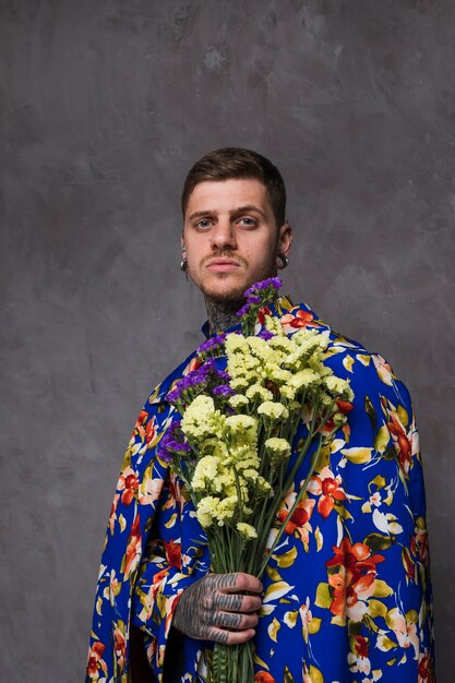 Het portret van een jonge mens met doordrongen oren en neus die gele en purpere limonium houden bloeit tegen grijze achtergrond