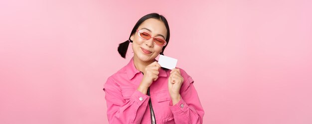 Het portret van een stijlvol, modern aziatisch meisje toont een kortingscreditcard en ziet er tevreden uit om contactloos winkelen te betalen op een roze achtergrond