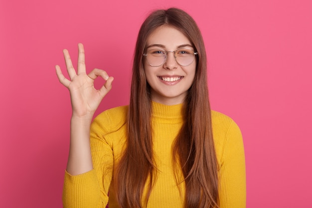 Het portret van gelukkig jong meisje met lang haar dat ok gebaar met haar vingers toont, kijkt glimlachend in goed humeur