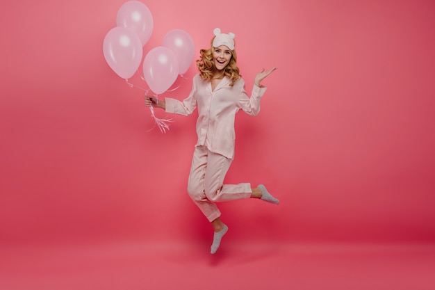 Het portret van gemiddelde lengte van aangenaam feestvarken in sokken die op roze muur springen. Leuke jonge vrouw in pyjama's en slaapmasker met plezier met helium ballonnen.