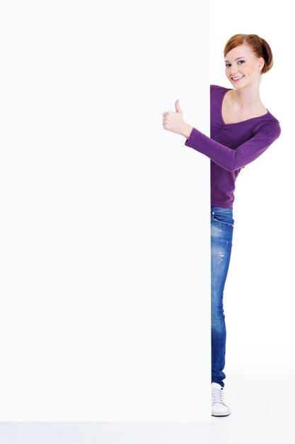 Het portret van gemiddelde lengte van een jonge vrouw kijkt uit wegens leeg aanplakbord met thumbs-up teken